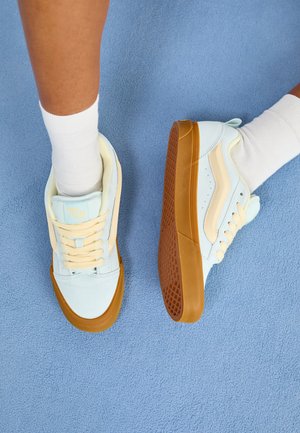 Zapatillas de color azul claro con acentos en crema, que presentan cordones, una suela de goma y un agarre texturizado. Llevadas con calcetines blancos sobre una superficie azul.