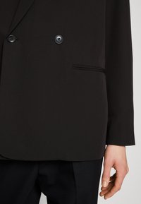 Blazer nero doppiopetto con una superficie liscia, caratterizzato da due bottoni tondi e una elegante tasca laterale con un sottile dettaglio.