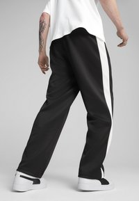 Sorte træningsbukser med en hvid side-stripe, lavet af glat materiale, parret med hvide chunky sneakers. Tatovering synlig på armen.