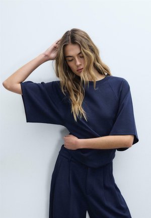 Jeune femme aux cheveux ondulés lâchés, portant un haut bleu marine ample à manches courtes et un pantalon large, debout avec un bras levé et les yeux fermés.