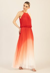 Rotes, plissiertes Kleid mit Halspartie, das nahtlos von tiefrot zu weiß übergeht. Das Modell trägt silberne High Heels.