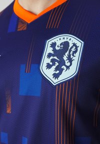 Maillot de football violet avec col orange, présentant un tissu texturé, des rayures diagonales, et un logo en forme de bouclier bleu clair avec un lion.