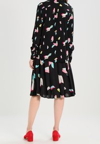 Vestido negro con patrones coloridos en rosa, azul y amarillo, con mangas largas y falda plisada, combinado con zapatos de tacón bloque rojos.