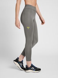 Grå högmidjade leggings med en mjuk textur och en gul bi-logotyp på låret, tillsammans med svarta och blåa träningsskor.