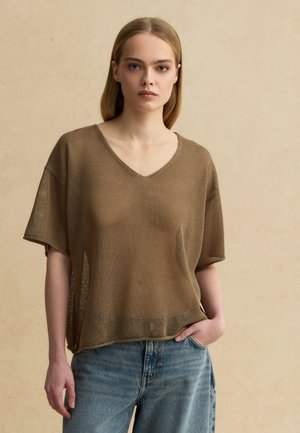 Joven con el pelo liso que lleva un suéter marrón suelto de punto y vaqueros azules, de pie con una mano en el bolsillo sobre un fondo liso.