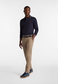 Marineblaues Langarm-Polohemd mit geripptem Kragen, beige Hose und marineblaue Turnschuhe mit Gummisohlen, stehend vor einem schlichten Hintergrund.