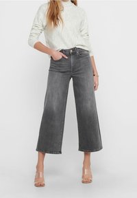 Nainen, joka on pukeutunut korkeavyötäröisiin, leveälahkeisiin harmaisiin farkkuihin, vaaleanharmaaseen pitkähiharaiseen neuleeseen ja läpinäkyviin avonaisiin korkokenkiin yksivärisellä taustalla.