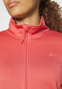 Veste zippée rouge avec un col montant, en tissu texturé, et un petit logo sur le côté gauche de la poitrine. Fini de surface lisse.