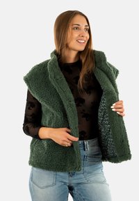 Gilet in pile verde con colletto alto, caratterizzato da una texture morbida e una fodera con stampa leopardata, indossato sopra una maglietta nera a maniche lunghe.