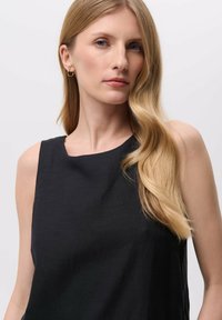 Zwarte mouwloze top met een ronde halslijn, gemaakt van lichte stof. Het model heeft lang, golvend blond haar en draagt kleine creolen.
