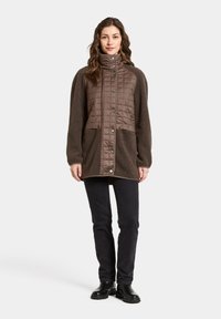 Bruine gewatteerde jas met fuzzy textuur, hoge kraag en drukknoopjes. Draagt over zwarte slim fit broek en zwarte laarzen.