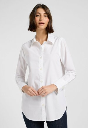 Camicia bianca con bottoni realizzata in morbido cotone, dotata di colletto appuntito, maniche lunghe e orlo curvo con sottili spacco laterale.