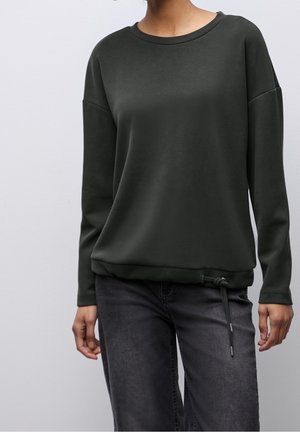 Personne portant un sweat-shirt à manches longues vert foncé avec un col rond et une taille ajustable, associé à un jean noir délavé.