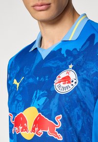 Maglia da calcio blu con motivo floreale, accenti gialli, colletto blu e loghi di FC Salisburgo e Red Bull.
