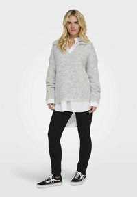 Jasnoszary, oversize sweter z dekoltem w serek i długimi rękawami, noszony na białej koszuli. W zestawie z czarnymi, dopasowanymi dżinsami i czarnymi sneakersami.