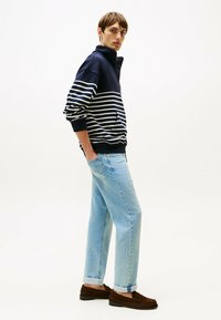 Felpa a righe blu navy e bianche con colletto alto, abbinata a jeans azzurri chiari e mocassini marroni; stile casual con vestibilità comoda.