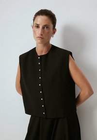 Svart ärmlös topp med rundad halsringning, framsidan har knappar i rad, strukturerad design och slät textur.