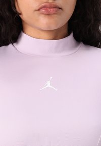 Világoslila, mock neck stílusú felső, puha anyagból készült, fehér Jordan logóval a mellen. A dizájn sima textúrájú és testhezálló fazonú.