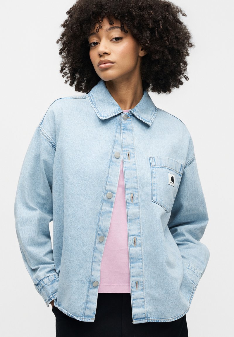 Light blue denim jacket with a collared neck, snap buttons, and a chest pocket featuring a logo patch. Worn over a pink shirt.