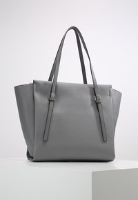 Sac fourre-tout structuré gris avec doubles bretelles d'épaule et surface texturée subtile, posé sur un piédestal blanc.