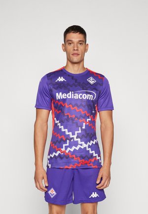 Uomo che indossa una maglia e pantaloncini sportivi viola con motivi a zigzag rossi, bianchi e neri e il logo dello sponsor "Mediacom", in piedi su sfondo bianco.