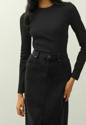 Frau trägt ein eng anliegendes schwarzes Langarmshirt, das in einen hoch taillierten schwarzen Jeansrock gesteckt ist, und steht vor einem schlichten Hintergrund.