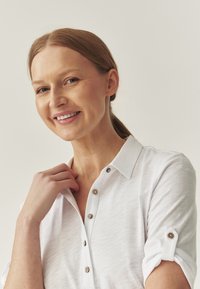 Biała koszula z kołnierzykiem, wykonana z teksturowanej tkaniny, z brązowymi guzikami i podwiniętymi rękawami. Modelka delikatnie dotyka dekoltu.