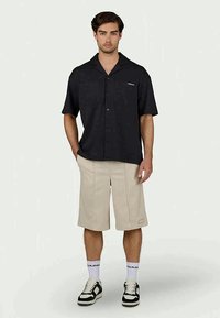 Camisa preta de manga curta com botões à frente e dois bolsos, combinada com calções bejes até ao joelho com pregas. Sapatos em preto e branco visíveis.