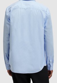 Uomo che indossa una camicia azzurra chiaro a maniche lunghe, con pantaloni neri, visto da dietro su uno sfondo semplice.