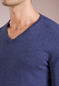 Pull bleu à col en V en tissu tricoté doux. Présente un petit logo brodé sur la poitrine gauche. Texture lisse avec une coupe ajustée.