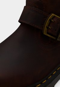 Σκούρο καφέ δερμάτινο παπούτσι με ραμμένο λουράκι και ορειχάλκινο πόρπη, με κίτρινο ράψιμο κατά μήκος της μαύρης άκρης της σόλας.