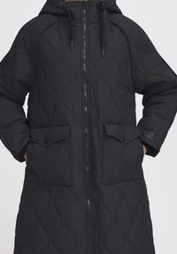 Sort quiltet lang frakke med h�ætte. Har en lynlås foran, to klaplommer og et tekstureret mønster af buede linjer på stoffet.