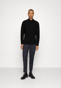Schwarze langärmelige Hemdbluse mit kragenähnlichem Saum, kombiniert mit dunkelgrauen maßgeschneiderten Hosen und schwarzen Schnürschuhen.
