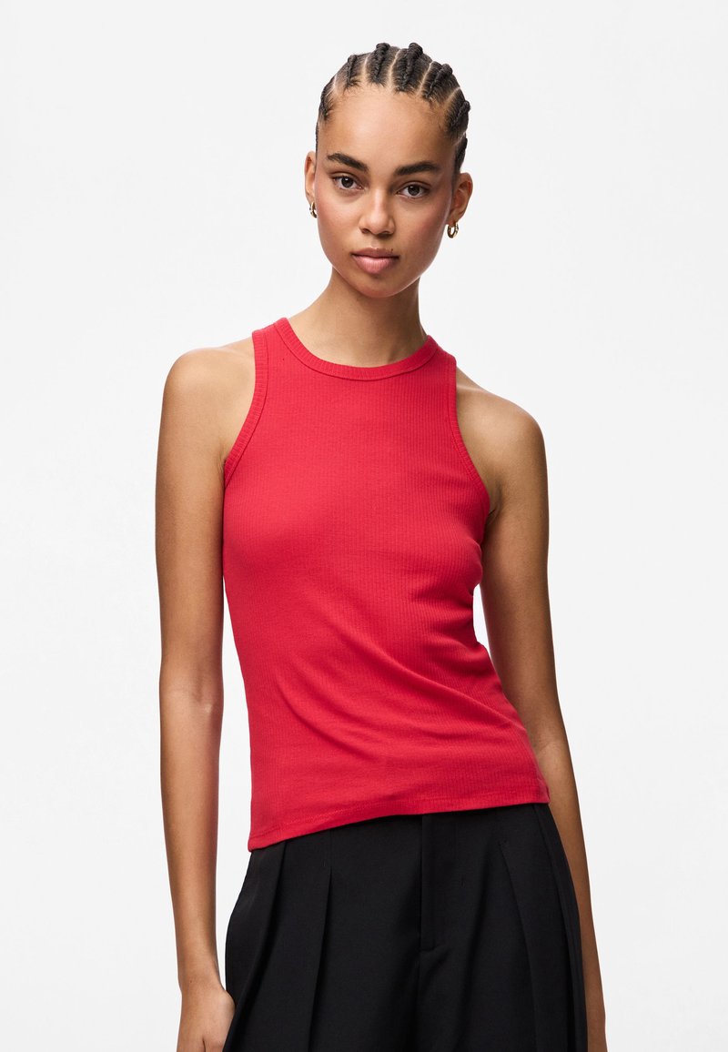 Kobieta z warkoczami nosząca złote kolczyki w kształcie kół, czerwoną bezrękawnikową bluzkę oraz czarne plisowane spodnie na jednolitym białym tle.