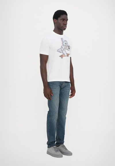 T-shirt blanche avec un graphisme de lapin cartoon coloré, associée à un jean bleu et des baskets grises. Le mannequin se tient devant un fond neutre.