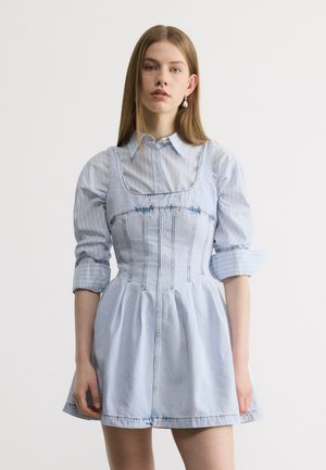 Fiatal nő egyenes hajjal, világoskék csíkos ingben, ujjatlan farmerruhára húzva, egyszerű fehér háttér előtt állva.