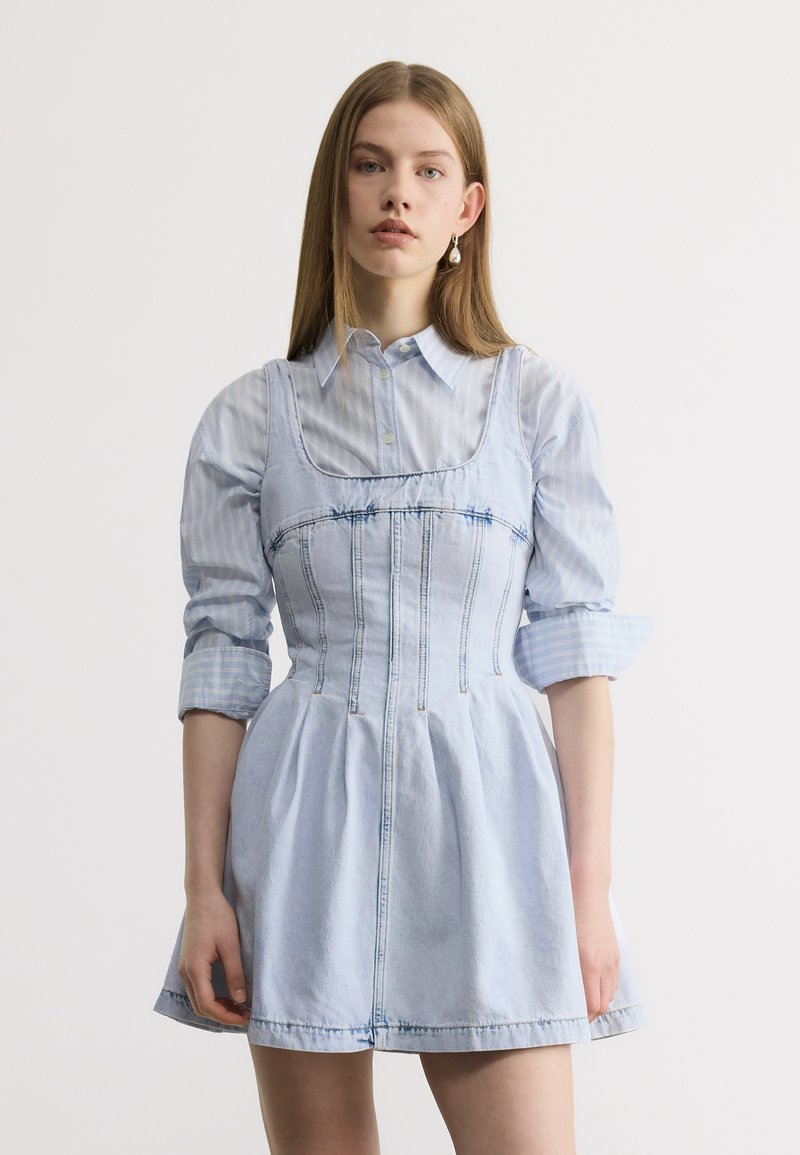 Νεαρή γυναίκα με ίσια μαλλιά, φοράει ένα ανοιχτό μπλε πουκάμισο με ρίγες κάτω από αμάνικο φόρεμα από τζιν, στέκεται μπροστά σε ένα απλό λευκό φόντο.