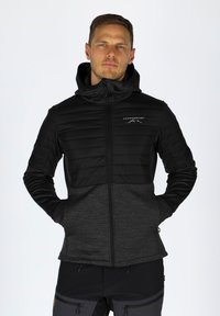 Giacca ibrida nera e grigia con zip frontale, parte superiore trapuntata e tessuto liscio. Presenta un cappuccio, tasche frontali e un logo sul petto.