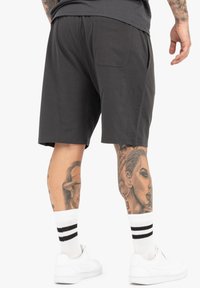 Graue Baumwollshorts mit einem elastischen Bund und Seitentaschen. Weiße Schuhe mit schwarzen Streifen sind sichtbar, neben tätowierten Beinen.