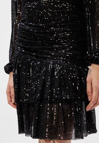 Vestido preto de lantejoulas com detalhes franzidos e bainha de camadas de babados. Mangas longas com punhos elásticos, apresentando lantejoulas brilhantes e textura transparente.