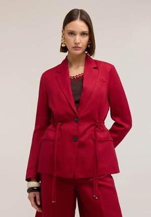 Kobieta nosząca czerwony, dopasowany blezer z regulowanym paskiem w talii oraz dopasowane spodnie, uzupełnione złotymi kolczykami i czarno-białymi bransoletkami.