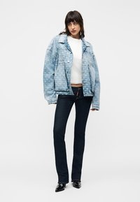 Femme portant une veste en jean bleu clair à motif surdimensionnée, un haut blanc, un jean droit foncé et des bottes noires à bout pointu, debout devant un fond blanc.