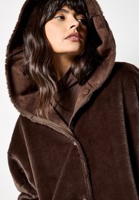 Cappotto in finta pelliccia marrone con un ampio cappuccio, caratterizzato da una texture liscia e chiusure a bottone sul davanti. Il design è oversize con bordi morbidi.