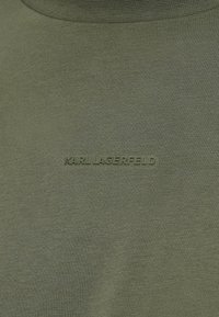 Olīvu zaļš krekls ar gludu tekstūru, ar paceltu, tonālu "KARL LAGERFELD" logotipu izšūtu priekšā.