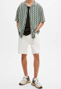 Chemise à manches courtes à motif géométrique en vert, noir et beige. Associée à un short blanc et des baskets multicolores.