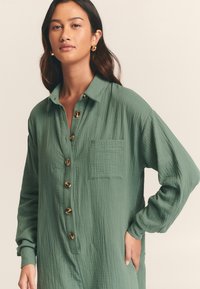 Zelená košeľa na gombíky vyrobená z textúrovanej tkaniny, s náprsním vreckom a veľkými okrúhlymi gombíkmi, s dlhými rukávmi a golierom.