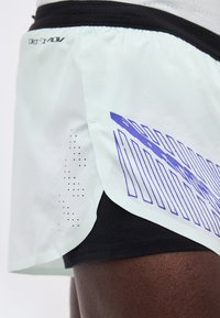 Αθλητικό σορτς ανοιχτού χρώματος με μαύρη εσωτερική επένδυση και μπλε αφηρημένη ρίγα στο πλάι, φορεμένα σε σκούρο δέρμα.