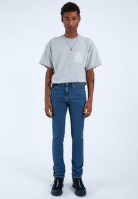 T-shirt grigia a maniche corte con un design stampato bianco sul petto, abbinata a jeans slim-fit blu e sneakers nere.