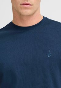 Tamsiai mėlynas trumpas rankovėmis medvilninis marškinėliai su apvaliu kaklu ir mažo siuvinėjimo logotipu ant krūtinės. Lygus paviršius, minimalistiškas dizainas.