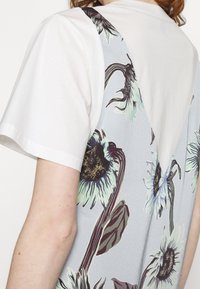 Gros plan d'une personne portant une chemise blanche à manches courtes sous un vêtement bleu clair avec un grand imprimé floral mettant en vedette des tournesols et des feuilles.